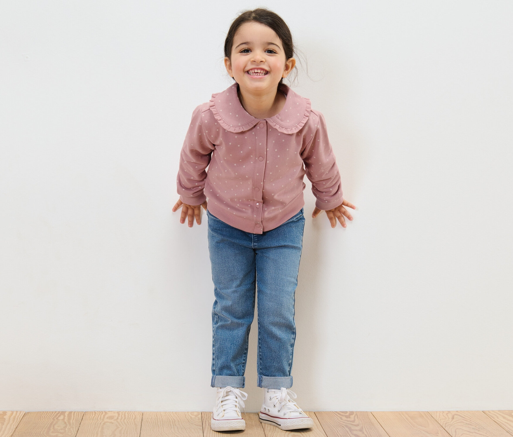 Usmívající se dívka stojí před bílou zdí a má na sobě džíny a dívčí mikinu s volánkem.