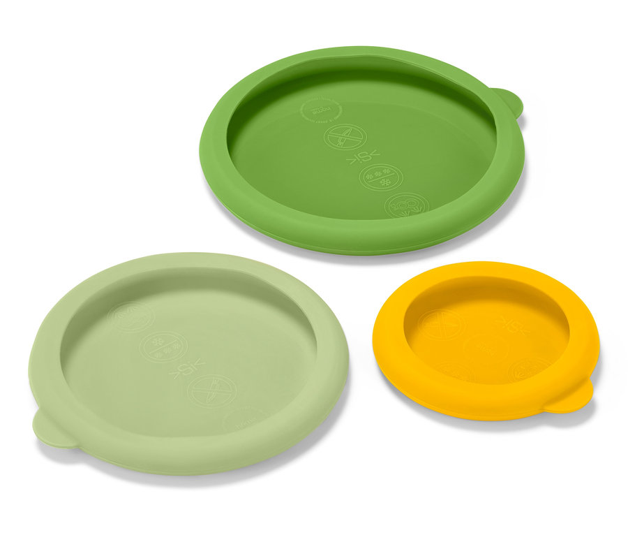 Tři silikonová víčka pro uchování čerstvosti, zelené, světle zelené a žluté, leží na bílém povrchu.