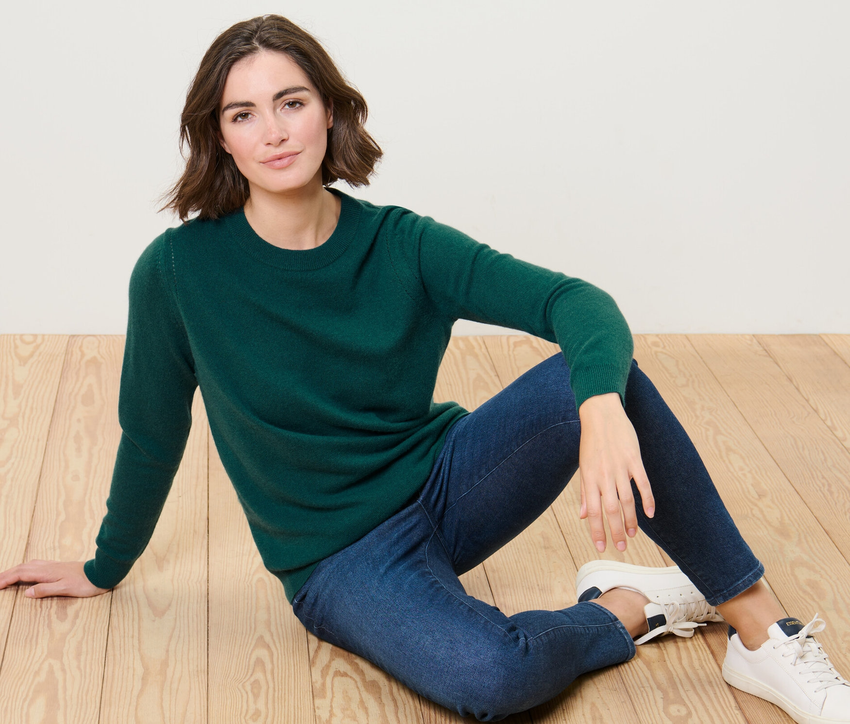 Žena sedí na podlaze v zeleném svetru z jemného úpletu s kašmírem a modrých džínách.