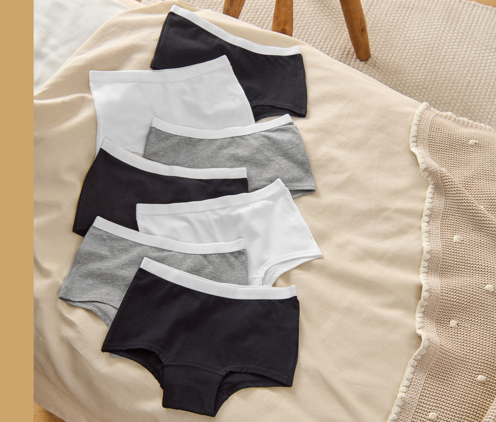Sedmery dívčí kalhotky (Dívčí kalhotky, 7 ks, šedá/bílá/černá) leží na béžové dece vedle dřevěné stoličky.