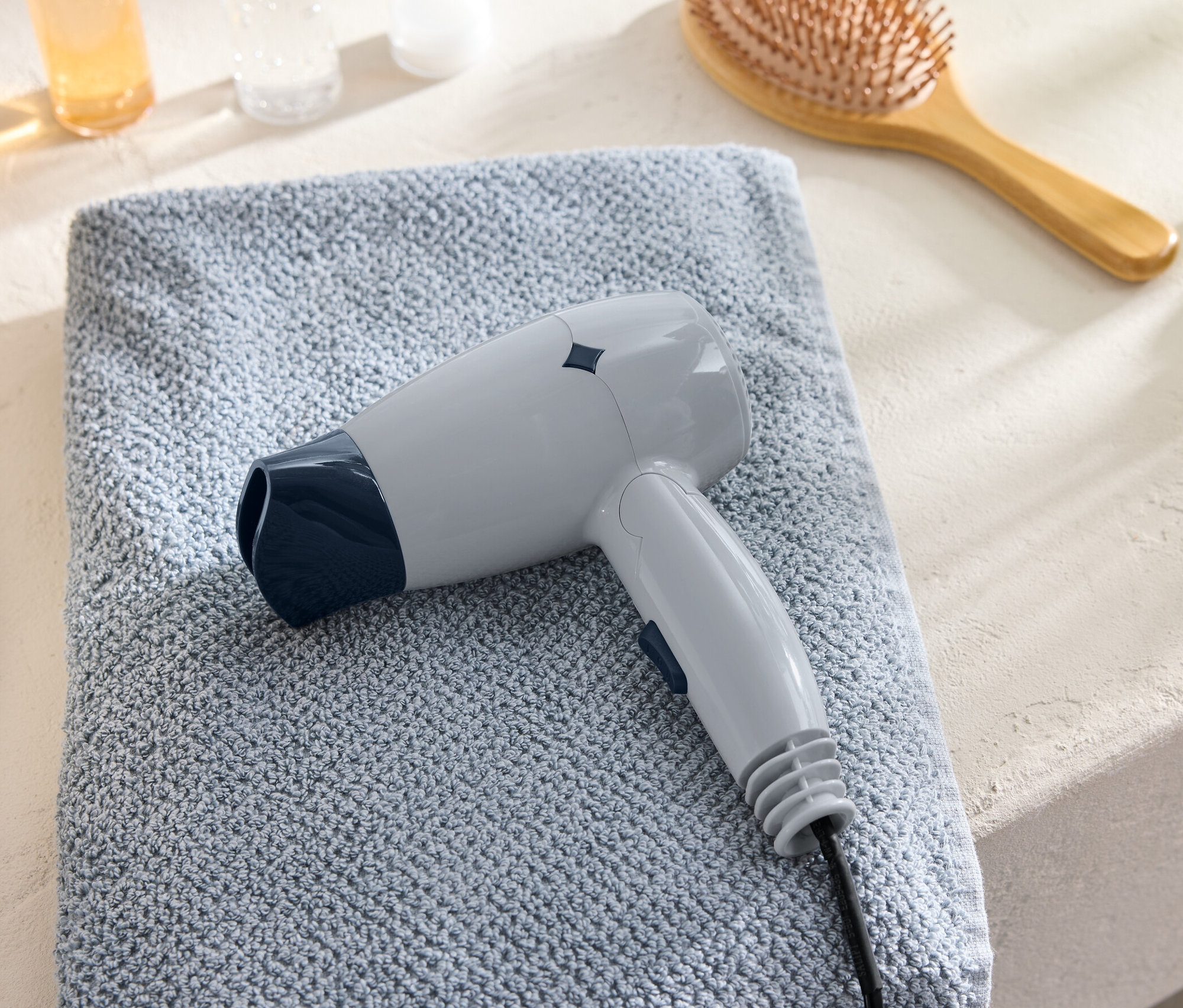Šedý cestovní vysoušeč vlasů leží na modrém ručníku vedle hřebenu.