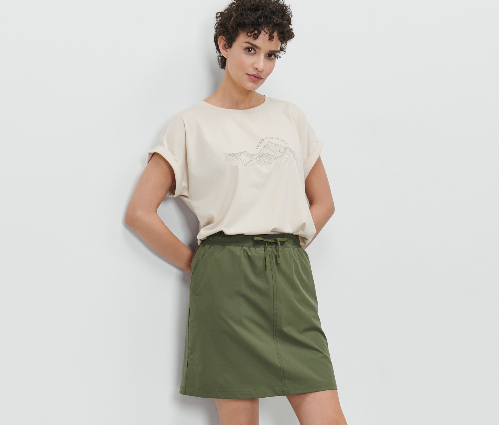 Žena pózuje v olivově zelené funkční sukni s kraťasy a béžovém tričku.