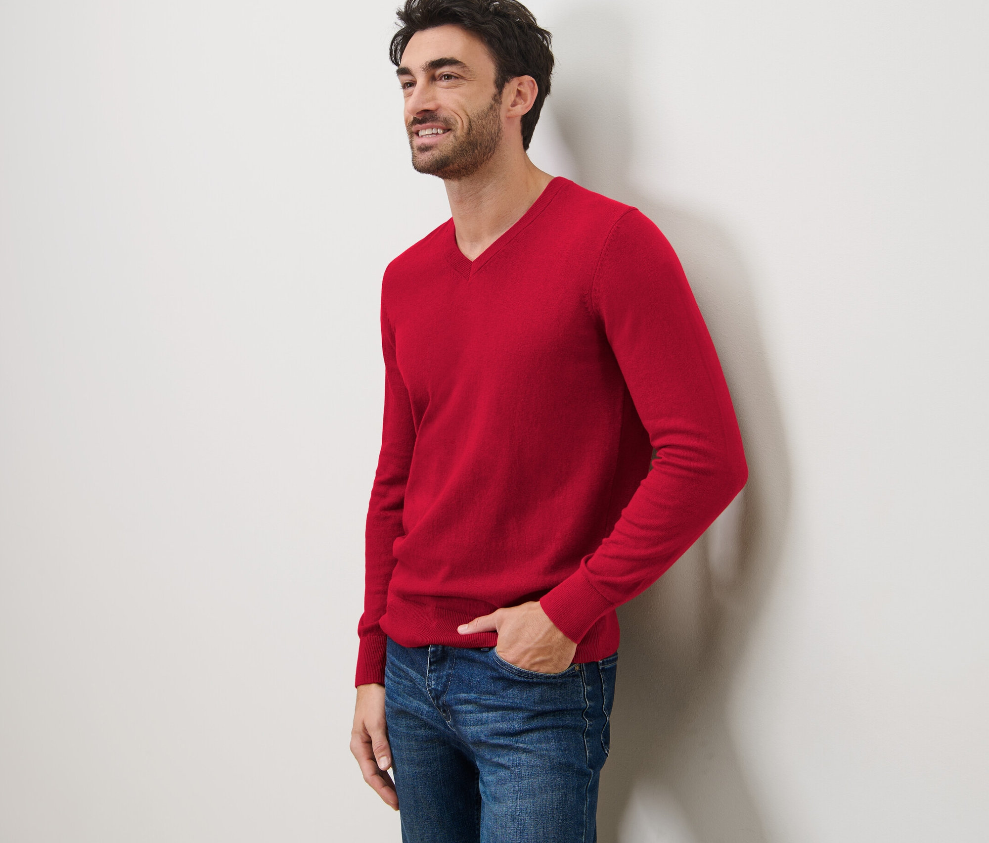 Muž se usmívá a opírá se o zeď. Má na sobě červený svetr s výstřihem do V a džíny.