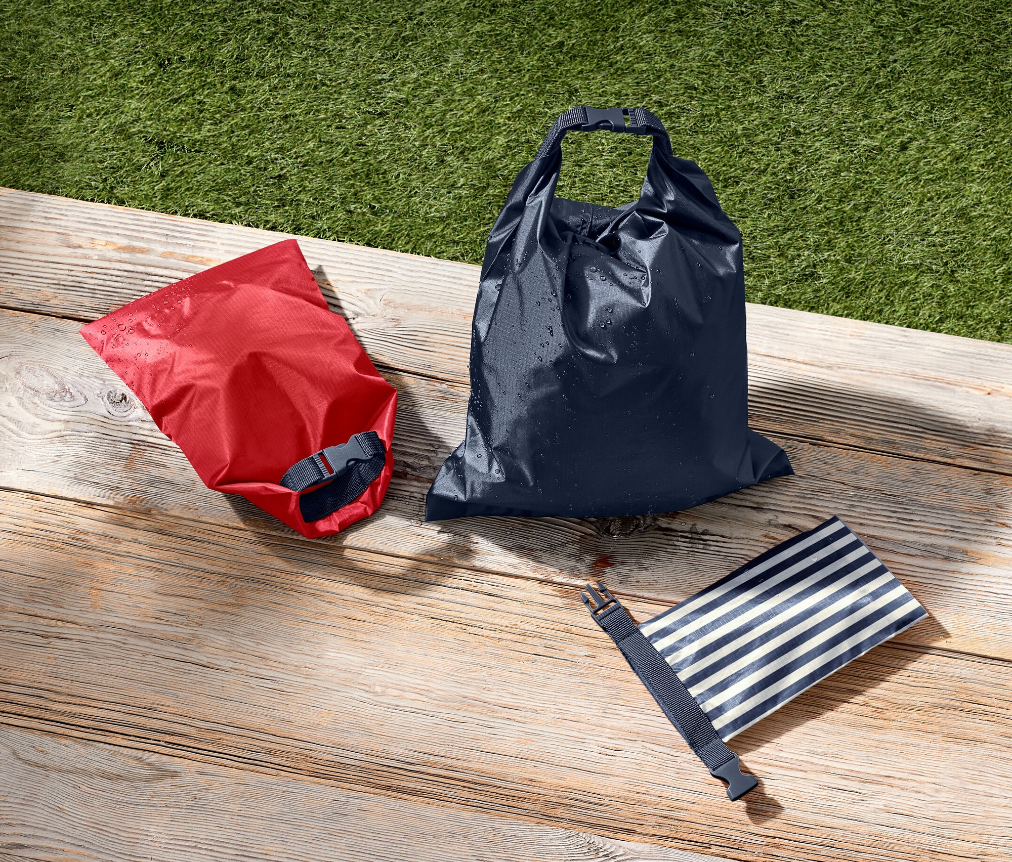 Červený, tmavě modrý a modro-bílý pruhovaný voděodolný vak leží na dřevěném povrchu vedle zelené trávy.