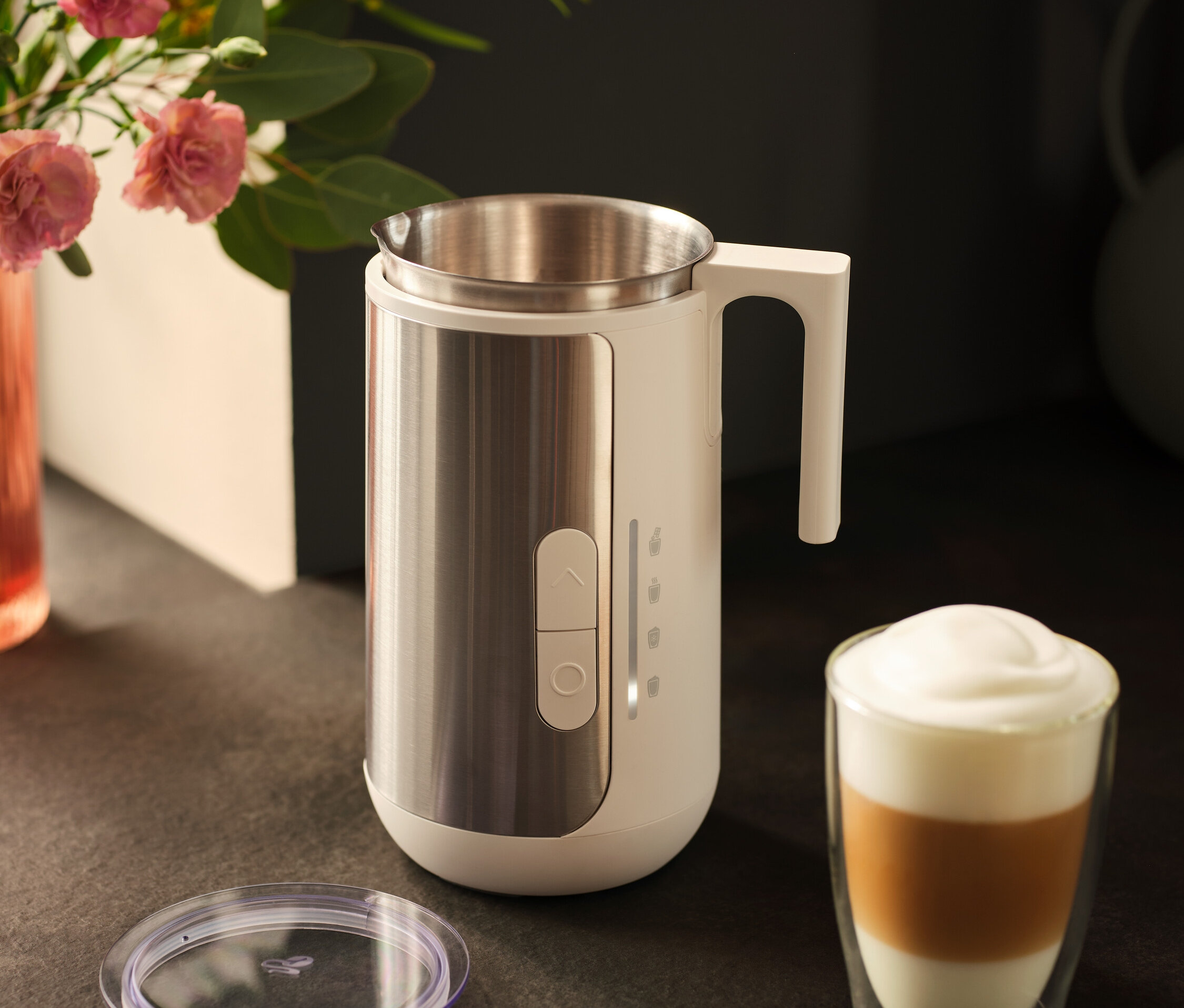 Nerezový napěňovač mléka stojí vedle sklenice s kávou latte a víka na tmavém pultu.