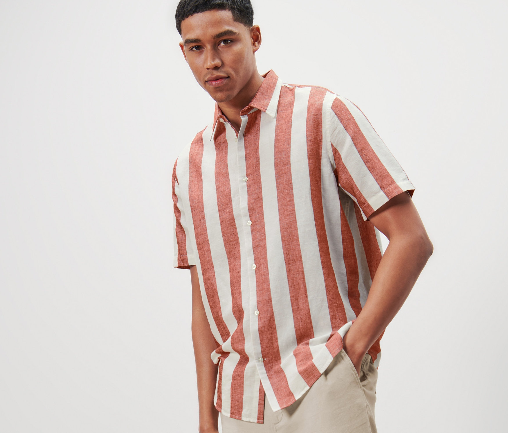 Muž pózuje v pruhované košili s krátkým rukávem s podílem lnu a světlých kalhotách.