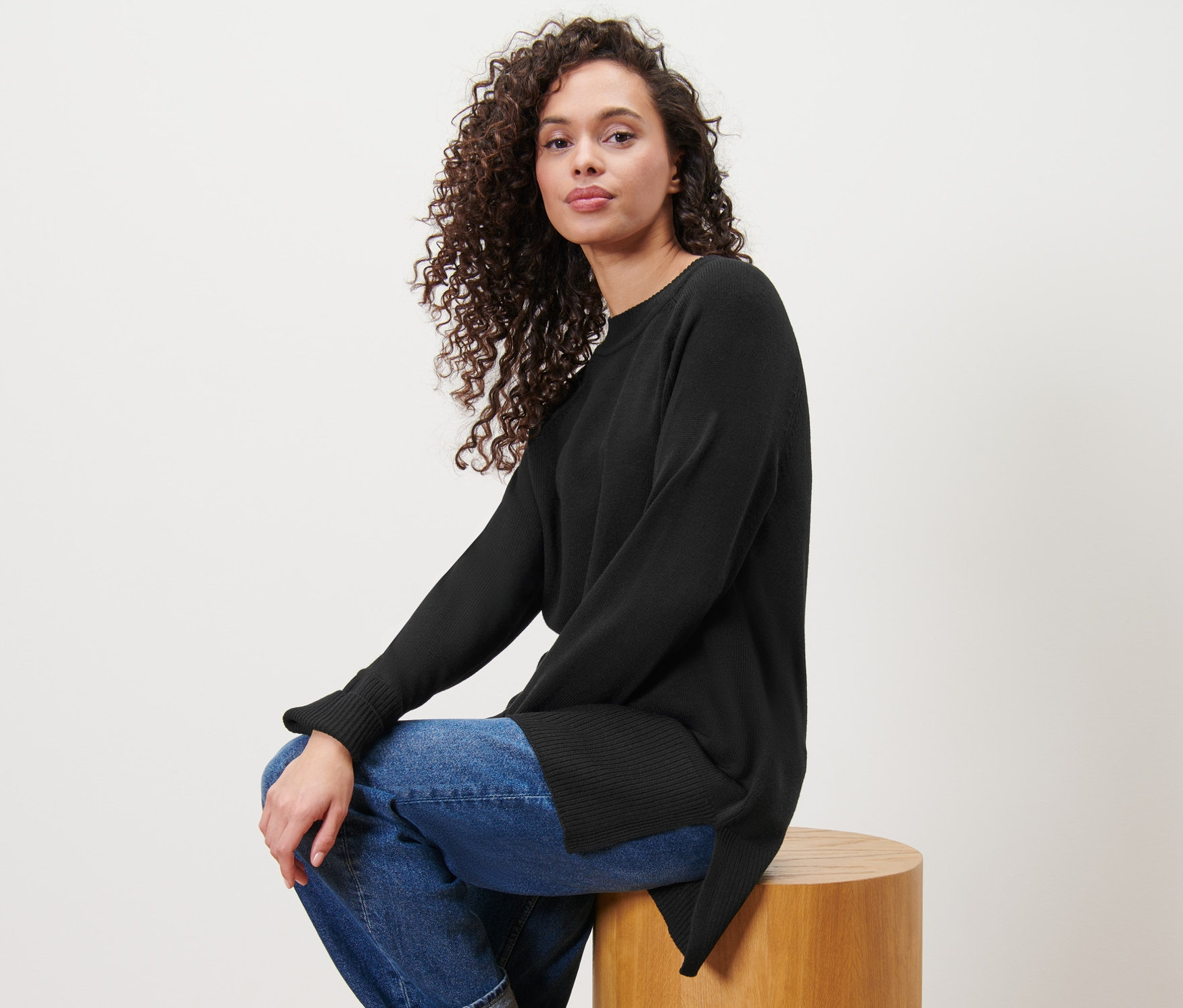 Žena sedí na dřevěné stoličce a má na sobě dlouhý černý pletený svetr.