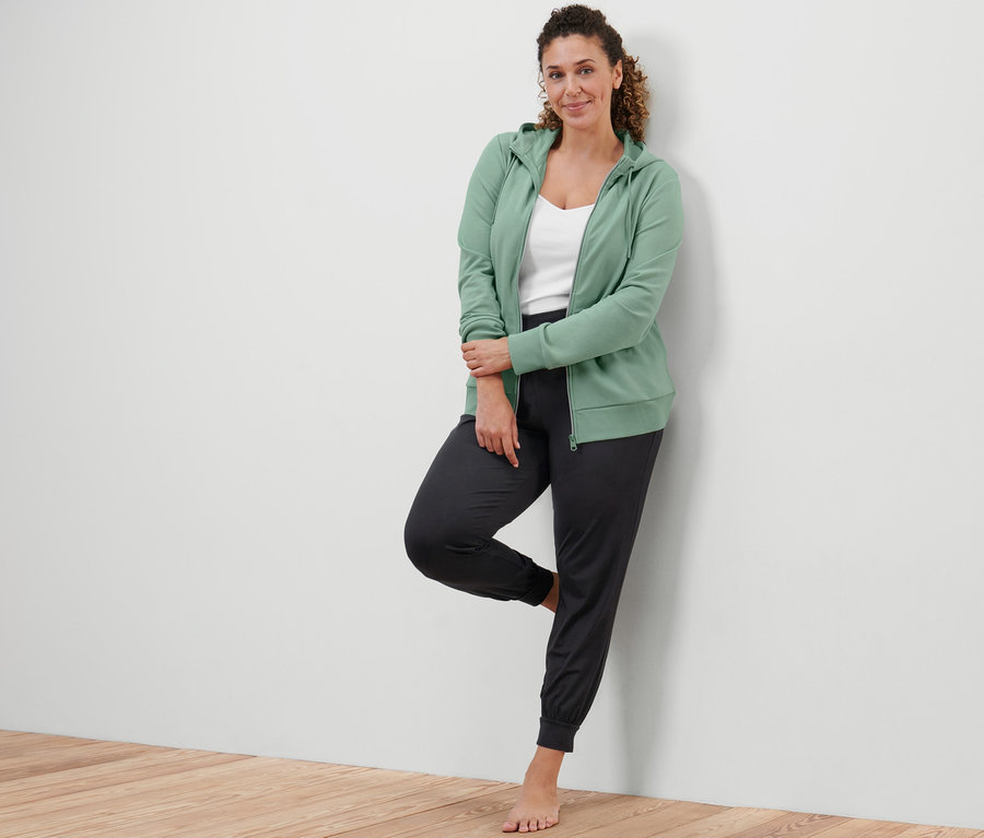 Žena opřená o zeď má na sobě šalvějově zelenou mikinu a kalhoty na jógu.