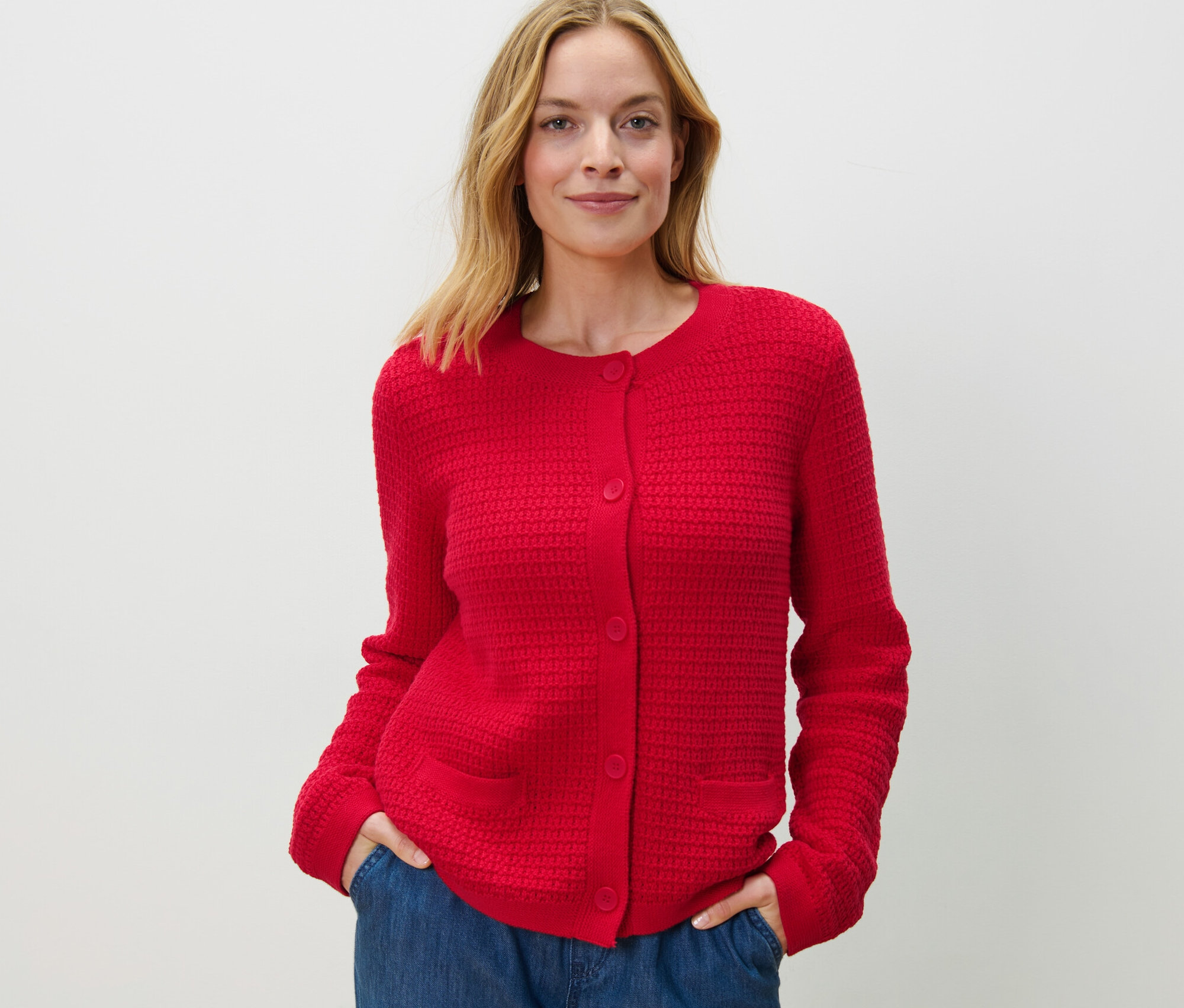 Žena pózuje v červeném kardiganu ze strukturovaného úpletu a modrých džínách.
