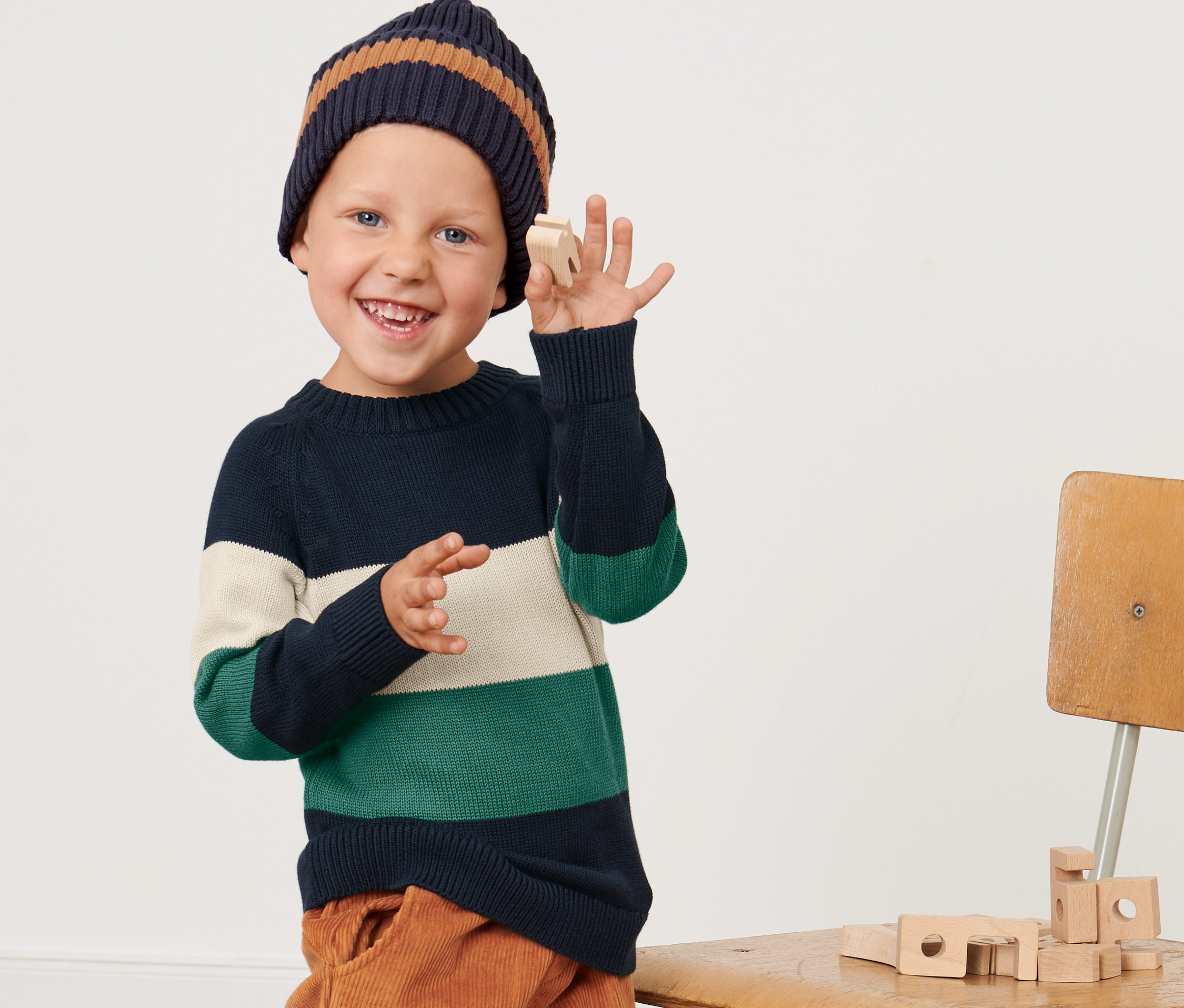 Chlapec v pleteném svetru přes hlavu a čepici drží dřevěnou hračku.