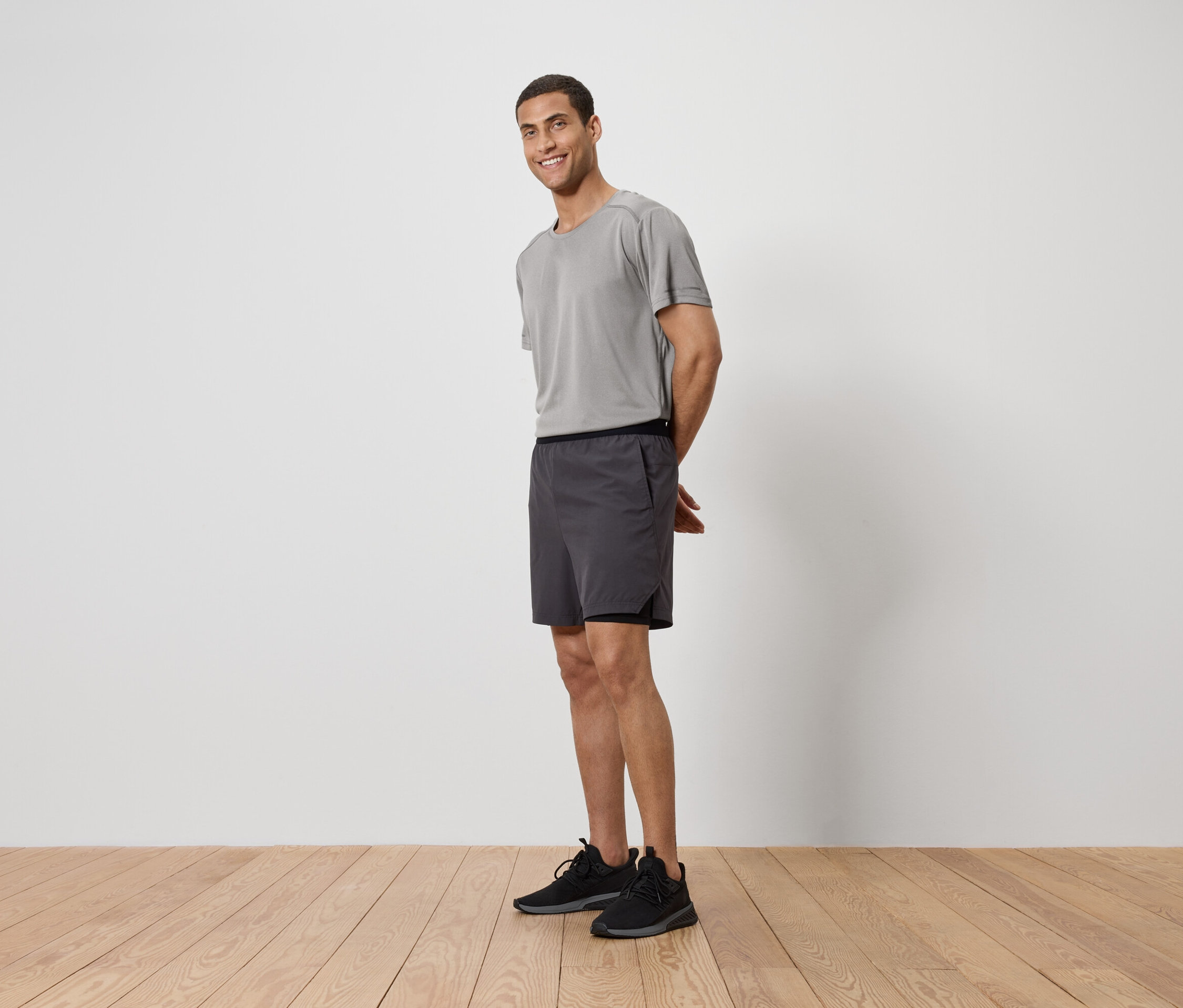 Muž v šedém tričku, šortkách a teniskách stojí na dřevěné podlaze před bílou zdí.