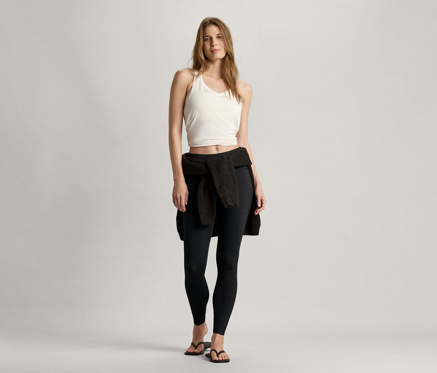 Žena stojí a má na sobě bílou bezešvou sportovní podprsenku, černé legíny a svetr uvázaný kolem pasu.