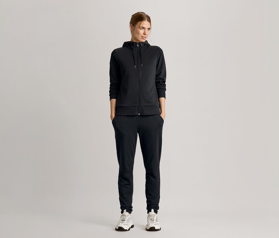 Žena stojí s rukama v kapsách, má na sobě černou teplákovou soupravu s mikinou s kapucí a tenisky.