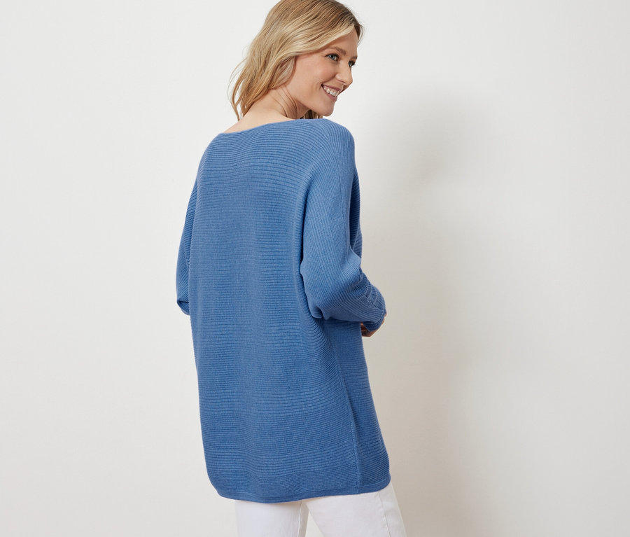 Blondýna se usmívá přes rameno a má na sobě modrý svetr a bílé kalhoty.