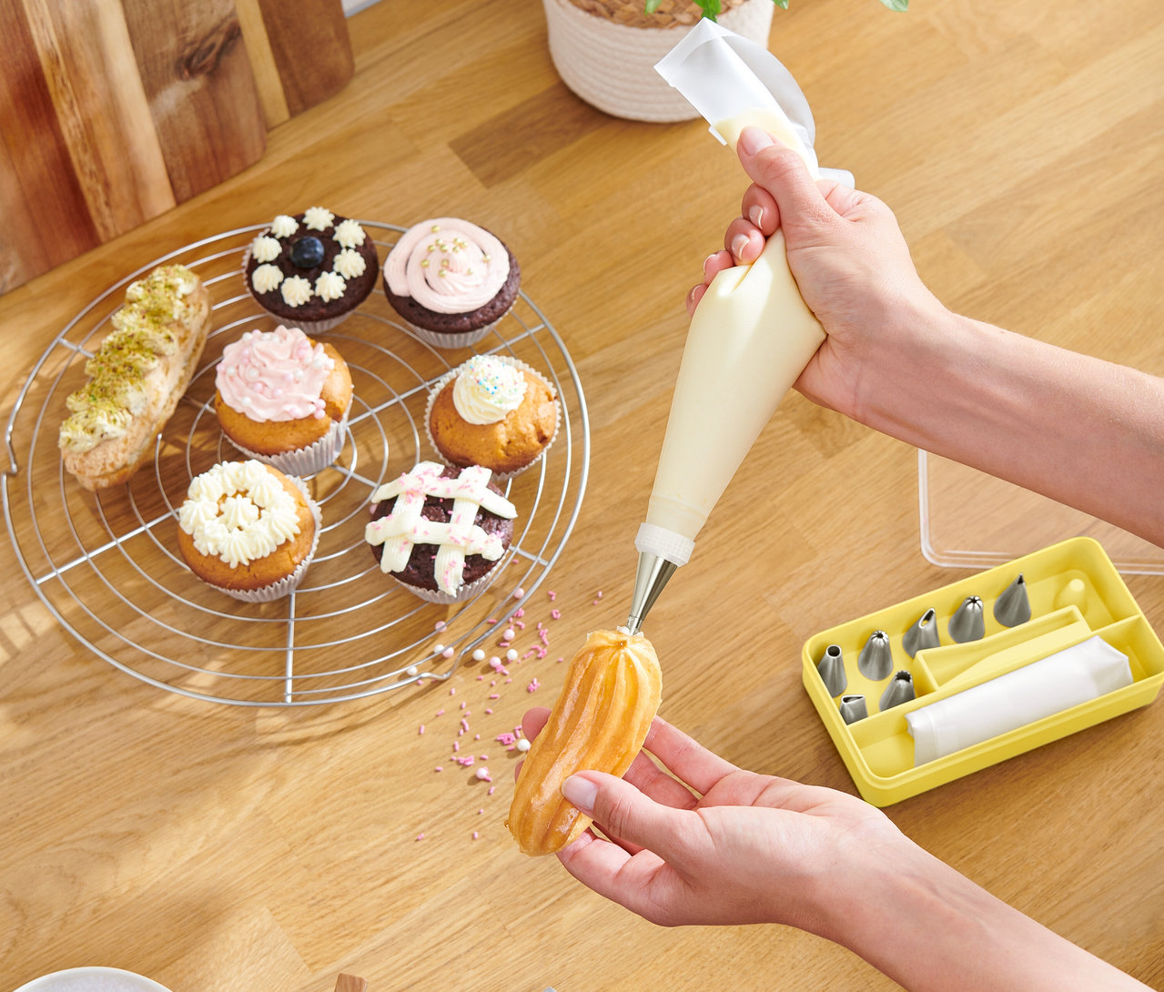 Osoba zdobí pečivo pomocí cukrářského sáčku, cupcakes a žlutá krabička s tryskami leží na dřevěném stole.