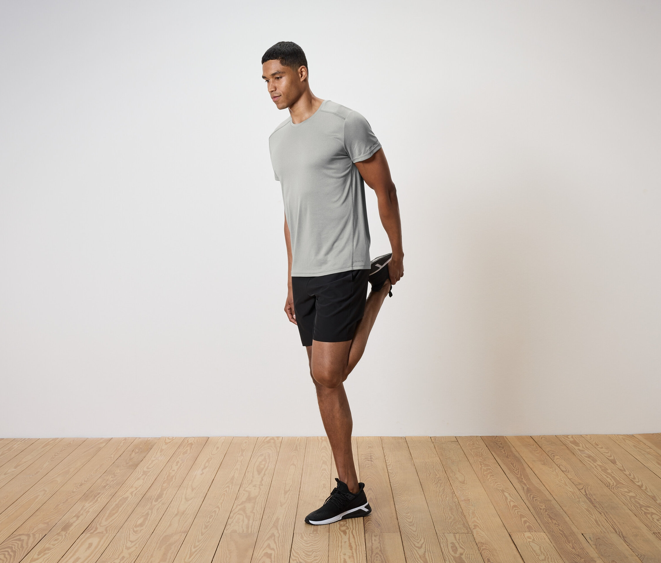 Muž v šedém tričku a černých šortkách protahuje nohu na dřevěné podlaze.
