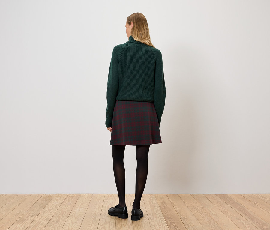 Žena stojí zády, má na sobě zelený svetr, kostkovanou sukni a černé punčocháče.