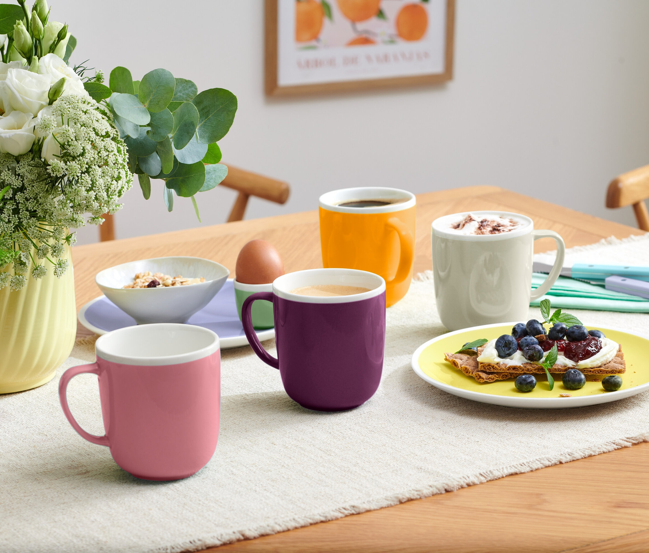 Na stole jsou čtyři hrnky různých barev, mísa s müsli, vejce v držáku a talíř s toustem s borůvkami.