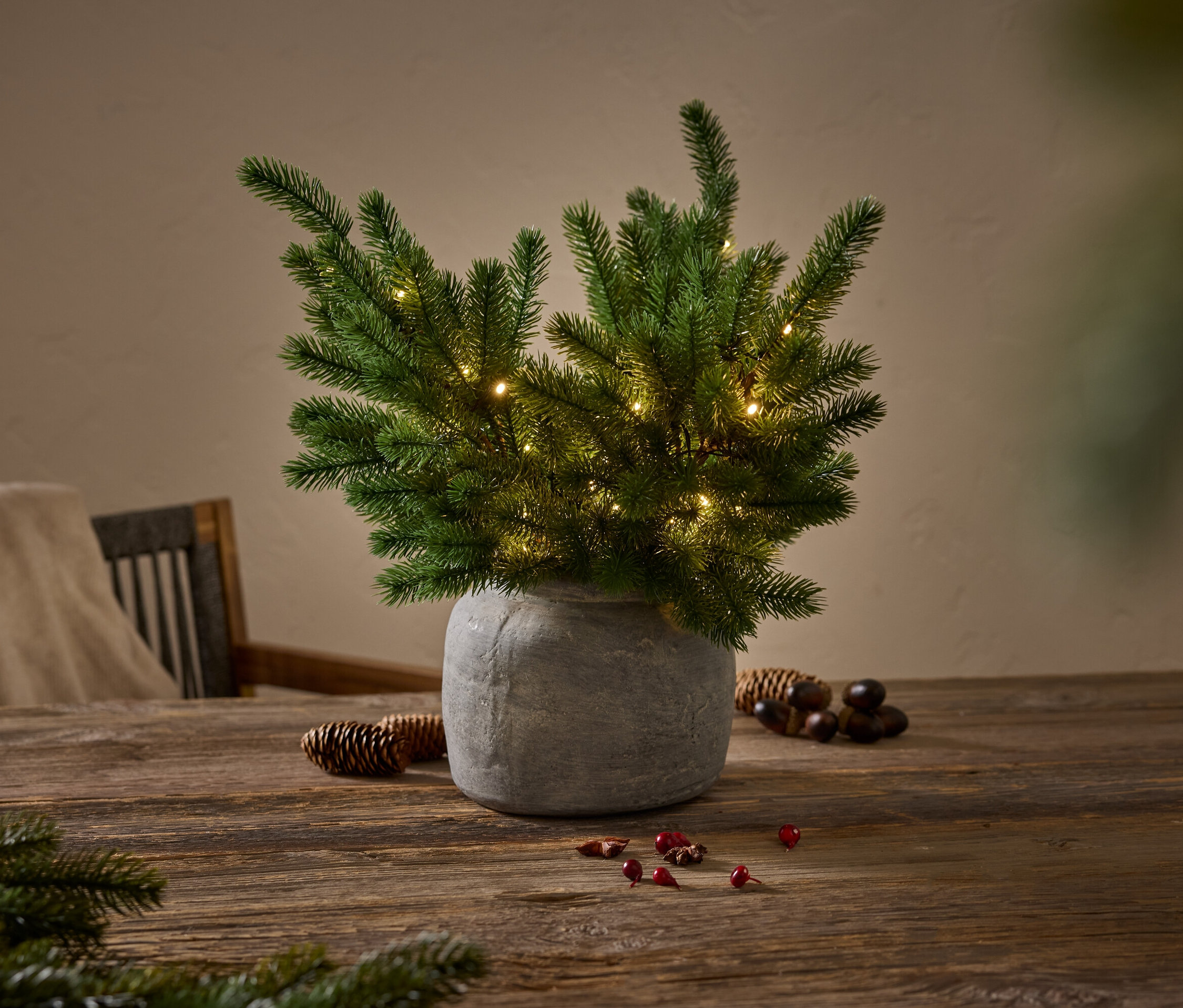 Zelený jehličnan s malými žárovkami v šedé váze na dřevěném stole.