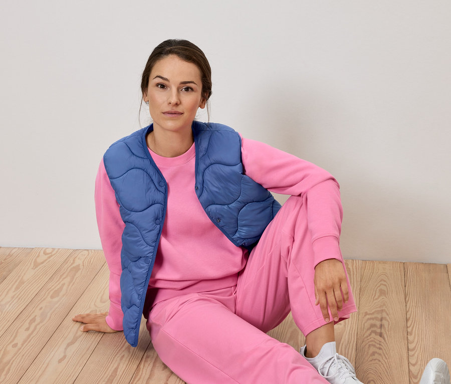 Žena sedí na podlaze v růžové soupravě a modré vestě.