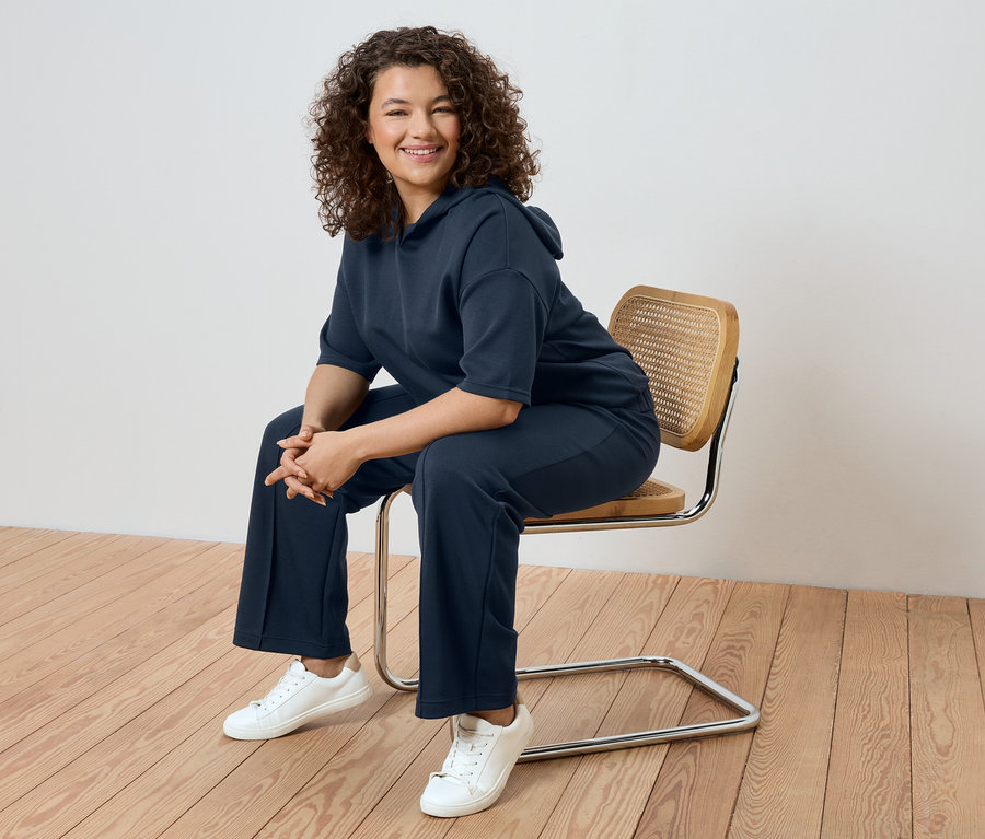 Žena s kudrnatými vlasy sedí na židli, oblečená v tmavě modré teplákové soupravě a bílých teniskách.