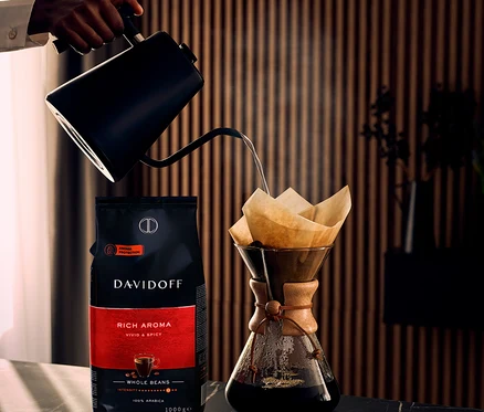 Ruka nalévá vodu z černé konvice s husím krkem do kávovaru Chemex vedle balení DAVIDOFF CAFÉ Rich Aroma - 3 x 1 kg Celá zrna.
