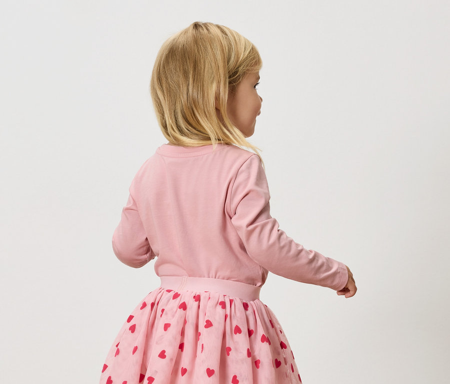 Malá blondýnka má na sobě růžové tričko s dlouhým rukávem a růžovou tylovou sukni s červenými srdíčky.