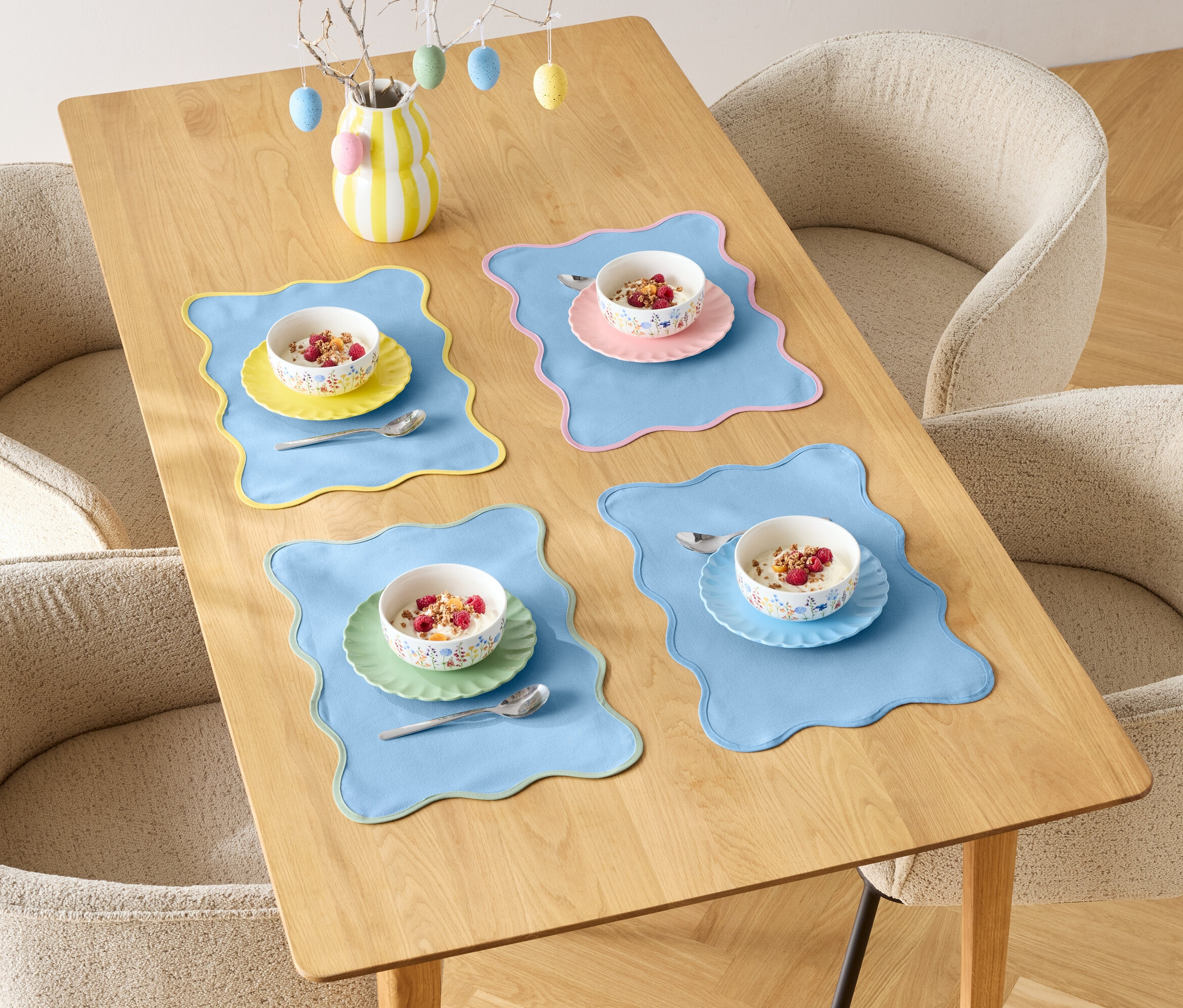 Jídelní stůl je prostřen pro čtyři osoby s miskami cereálií a barevnými prostíráními, obklopený čtyřmi židlemi.