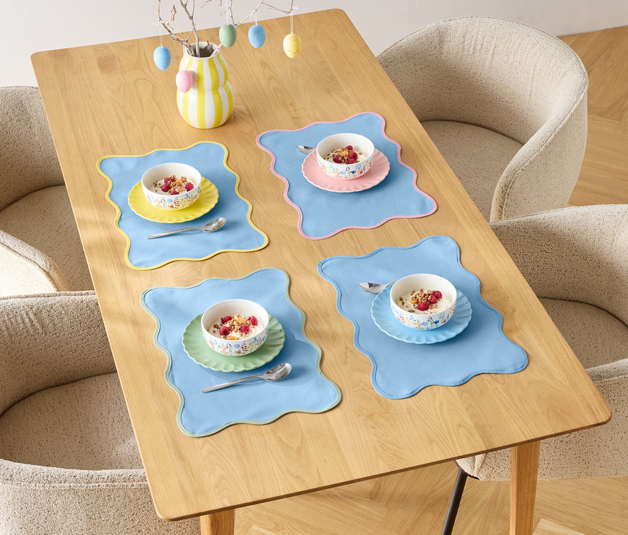 Jídelní stůl je prostřen pro čtyři osoby s miskami cereálií a barevnými prostíráními, obklopený čtyřmi židlemi.