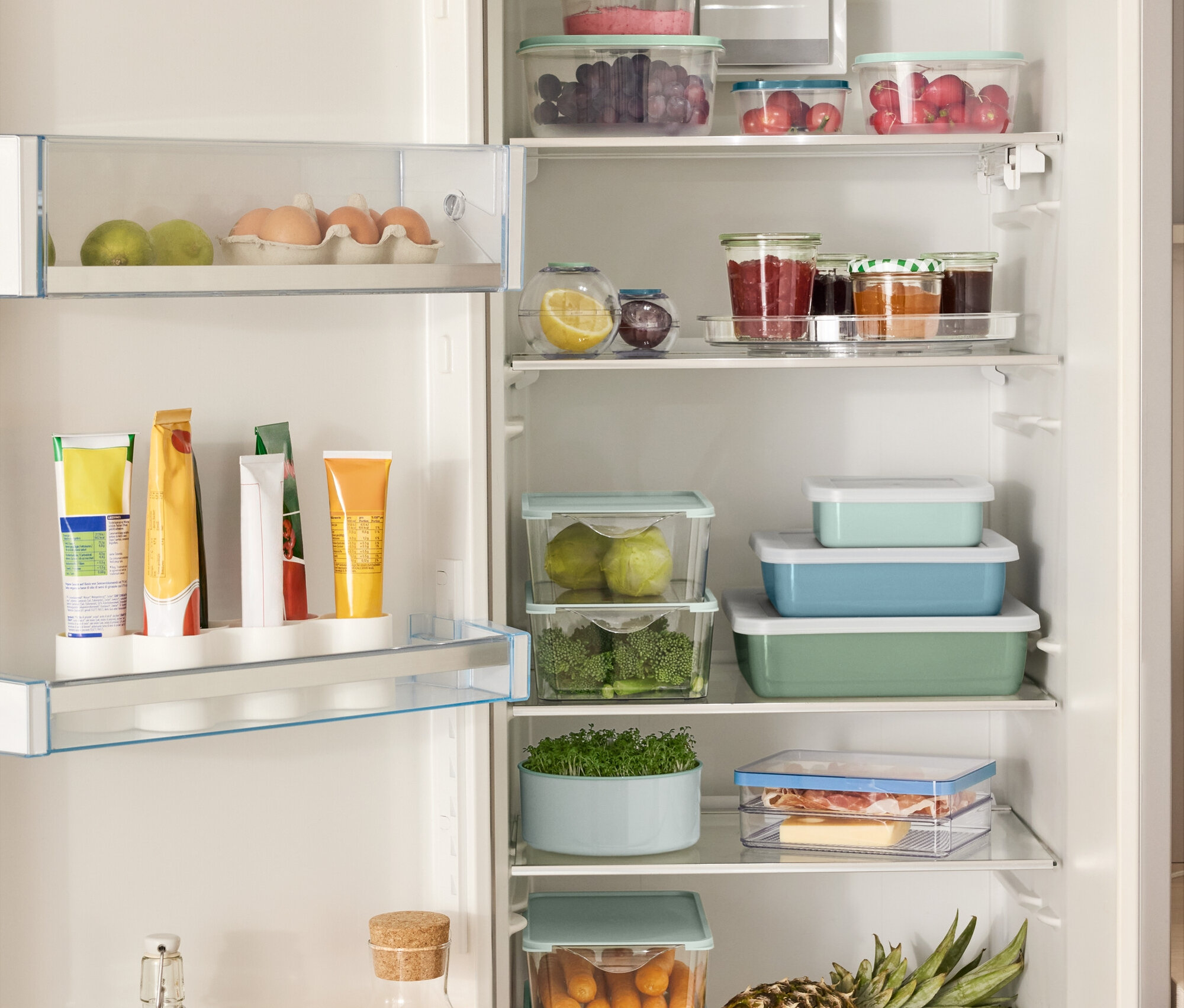 Otevřená lednice plná různých potravin, včetně 4 úložných boxů, 2 kulatých nádob na uchovávání potravin, 3 smaltovaných zapékacích forem s víkem a otočného talíře.