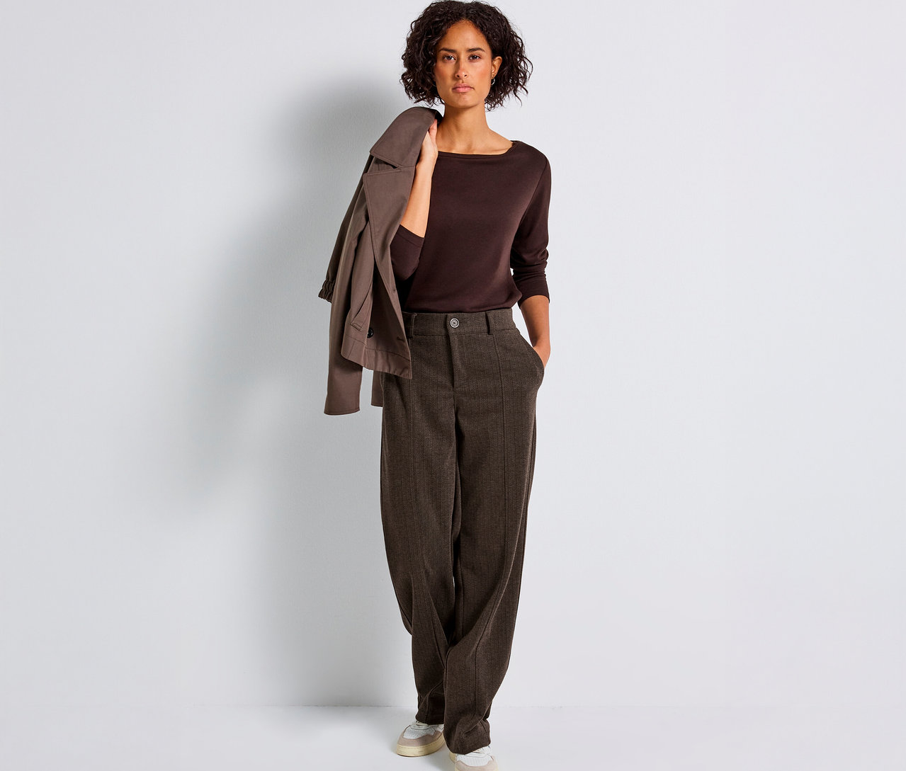 Žena stojí a má na sobě hnědé kalhoty s rovnými nohavicemi Street One, hnědý top a sako přes rameno.