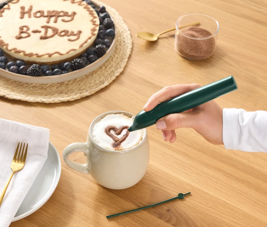 Ruka kreslí zeleným dekoračním perem kakaové srdce na pěnu kávy v hrnku. Na dřevěném stole je také vidět dort s nápisem „Happy B-Day“ a bobulemi, sklenice kakaového prášku, zlatá lžička a talíř s ubrouskem a vidličkou.