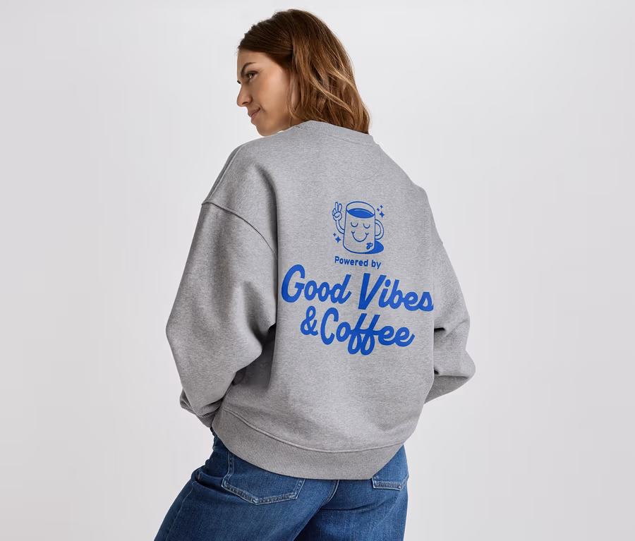 Žena má na sobě šedou mikinu s modrým nápisem "Good Vibes & Coffee" na zádech a džíny.