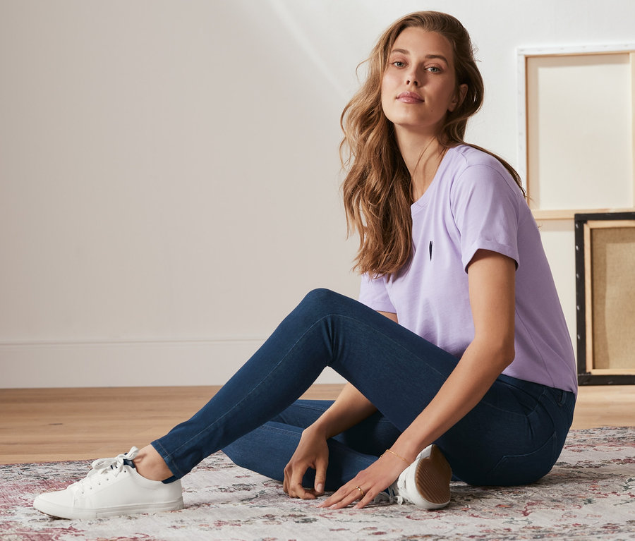 Žena sedí na koberci a má na sobě denimové džegíny středně modré barvy, fialové tričko a bílé tenisky.