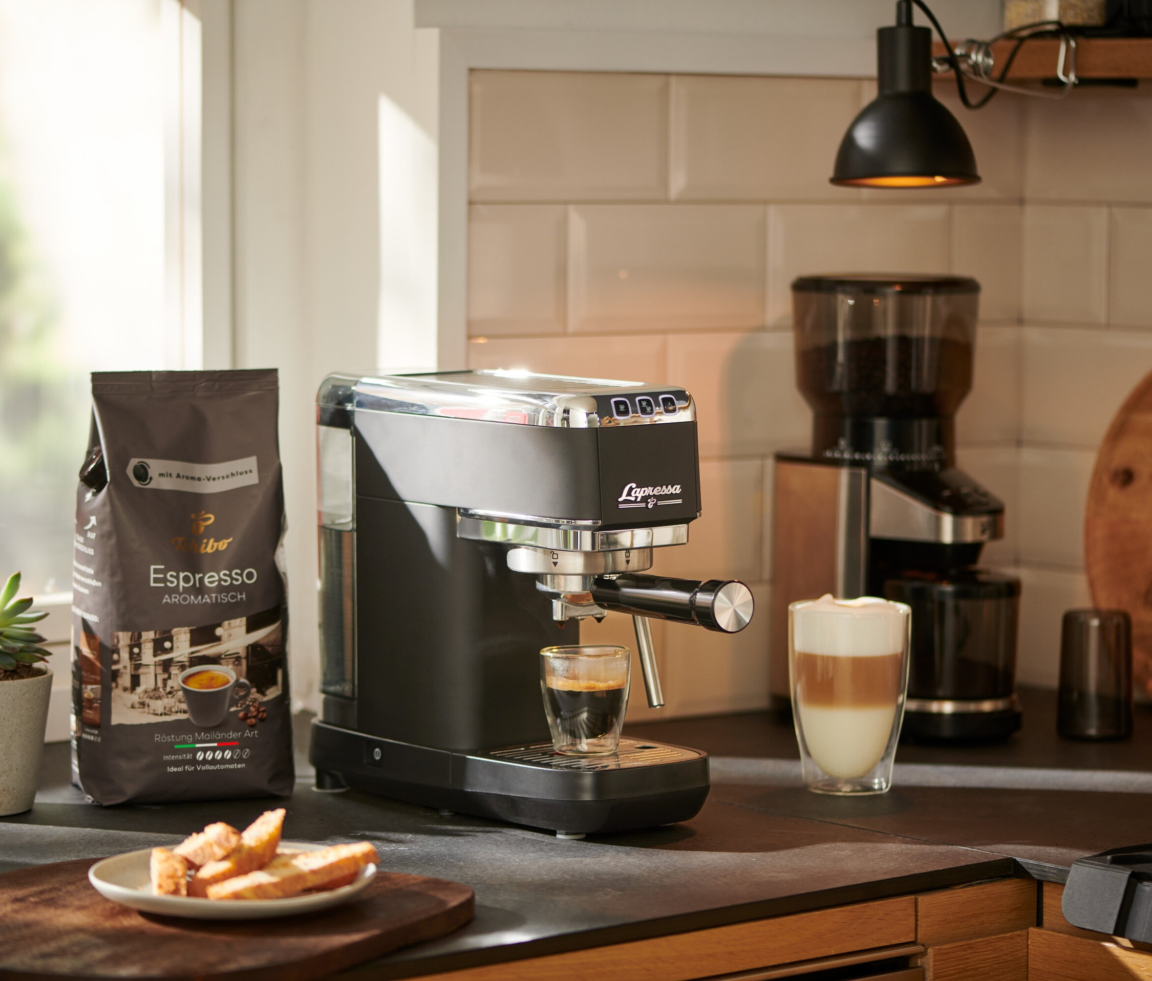 Espresso vytéká z pákového kávovaru »Lapressa«, černý, do sklenice. V pozadí je mlýnek na kávu a balíček kávy.