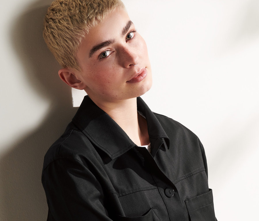 Mladá osoba s krátkými blond vlasy nosí černou košilovou bundu.