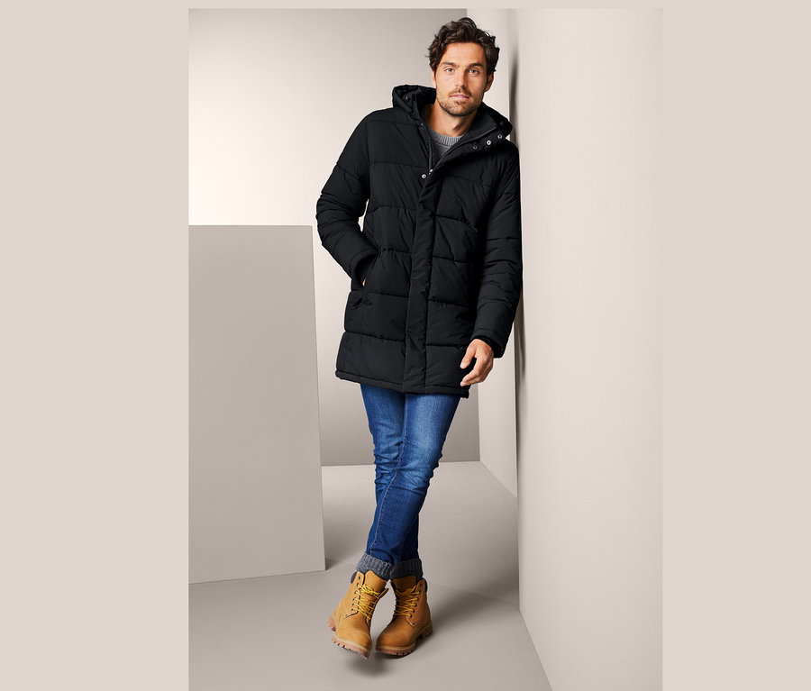 Muž opřený o zeď má na sobě prošívanou parku s kapucí, džíny a žluté boty.