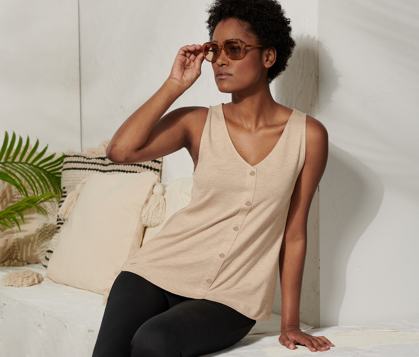 Žena sedí a upravuje si sluneční brýle. Má na sobě béžový top s knoflíkovou légou a černé legíny.