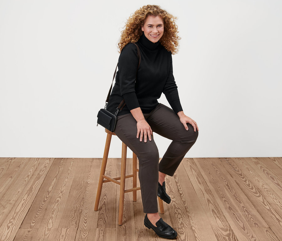 Žena sedí na stoličce a má na sobě černý rolák z jemného úpletu, hnědé bengalínové kalhoty, kožené mokasíny a minikabelku.