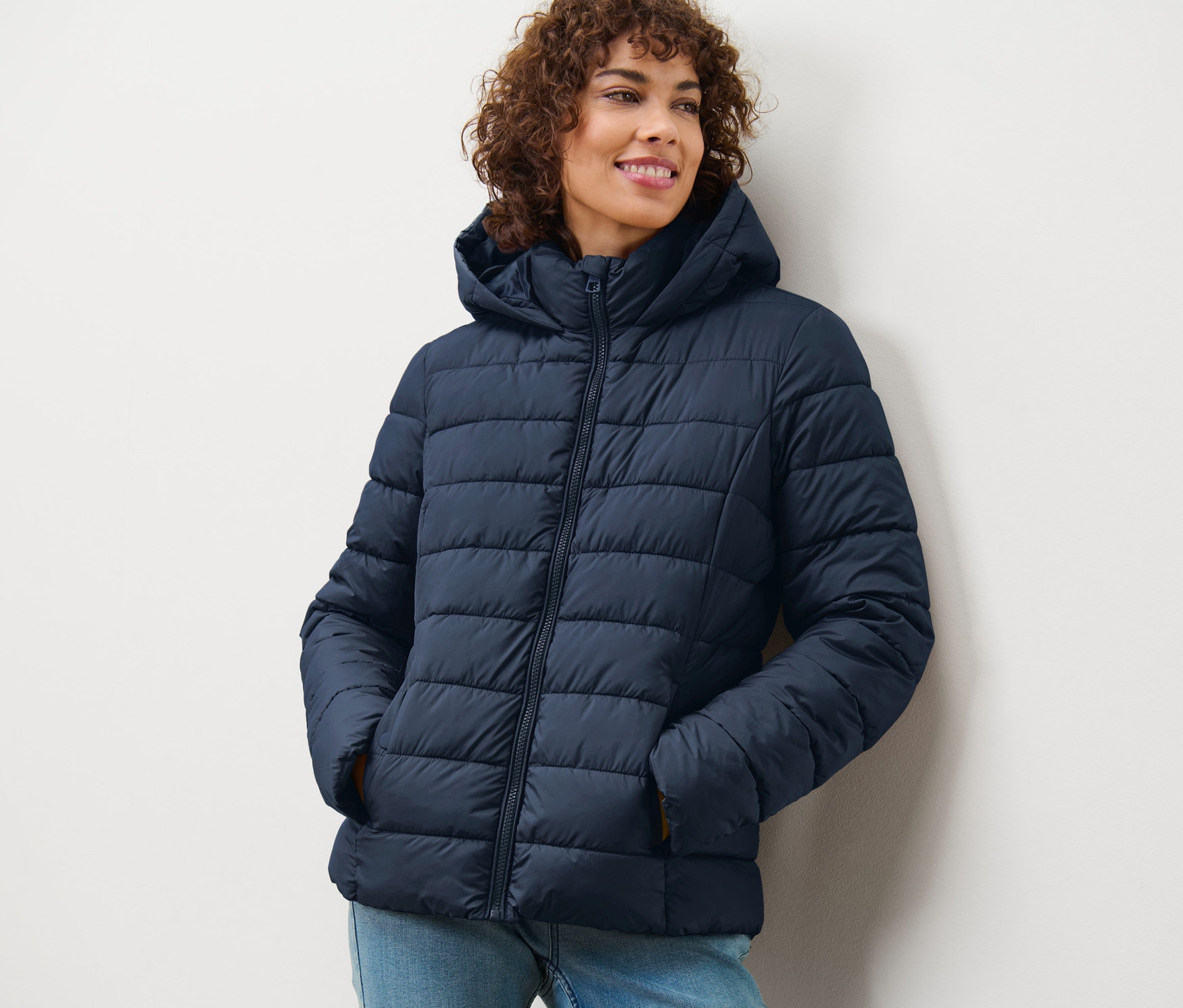 Žena usmívá se a má na sobě tmavě modrou prošívanou bundu s kapucí.