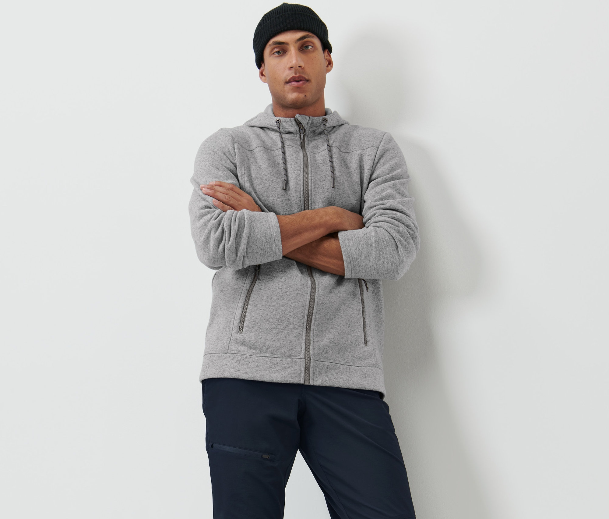 Muž se zkříženýma rukama pózuje v šedé bundě z pleteného fleecu a černé čepici.