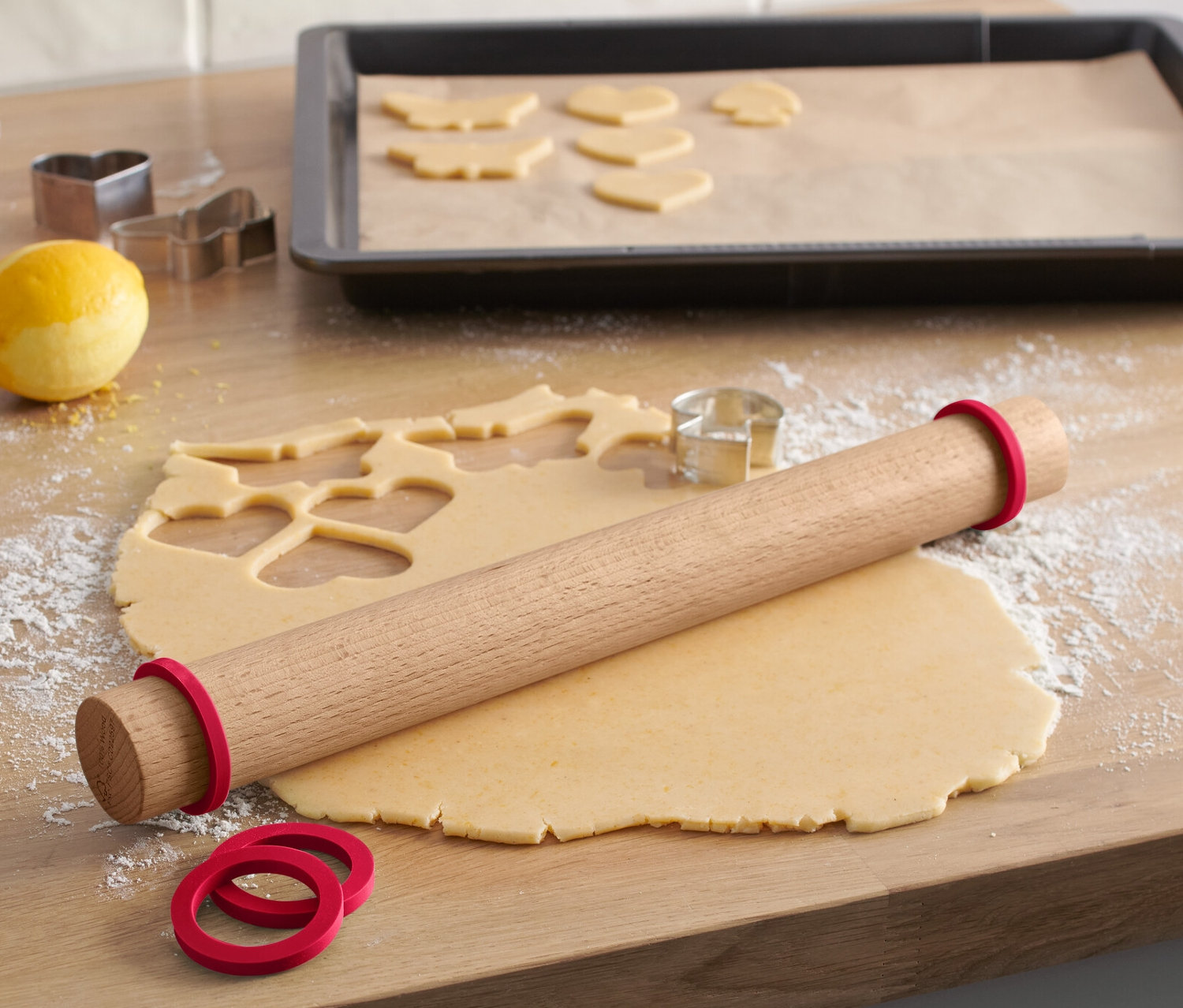 Na dřevěné desce leží dřevěný váleček na těsto s červenými gumičkami, vedle je rozválené těsto s vykrájenými tvary a formičky na vykrajování.