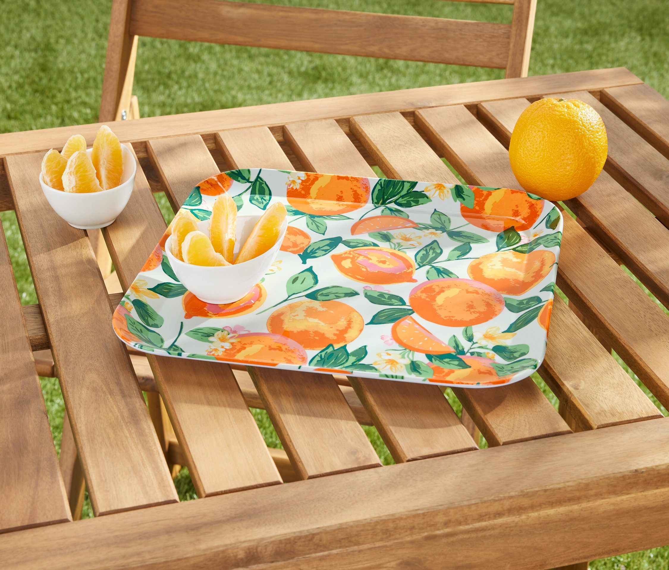 Na dřevěném stole stojí podnos s motivem pomerančů, miska s měsíčky pomeranče a celý pomeranč.