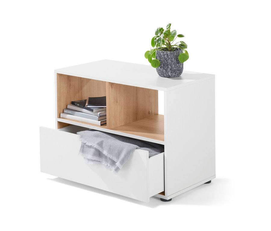 Bílá nízká skříňka se zásuvkou, na které je položená rostlina v šedém květináči. V otevřené přihrádce jsou knihy a v zásuvce šedá deka.