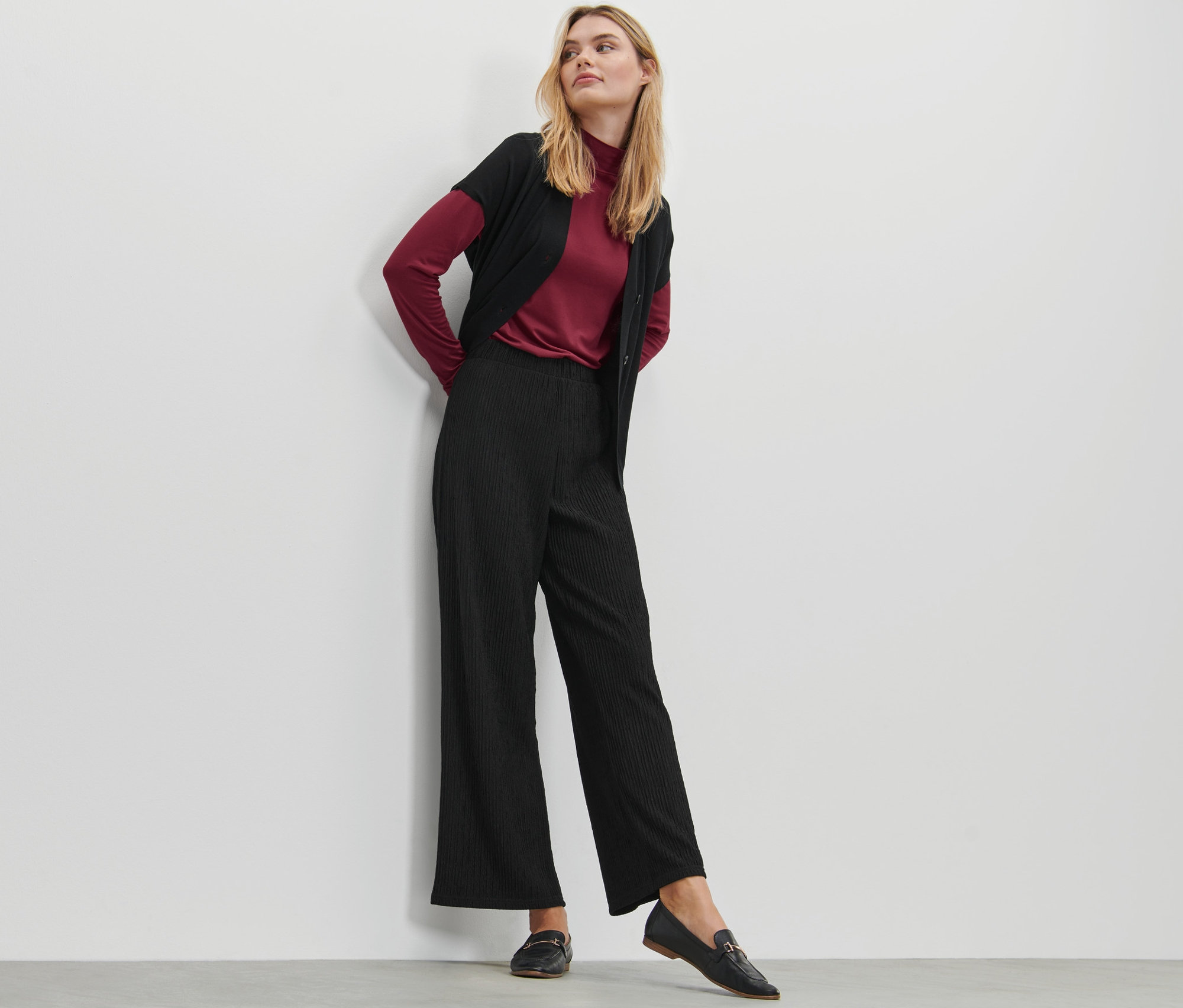 Blondýna pózuje v černých plisovaných kalhotách, vínovém roláku a černém svetru.