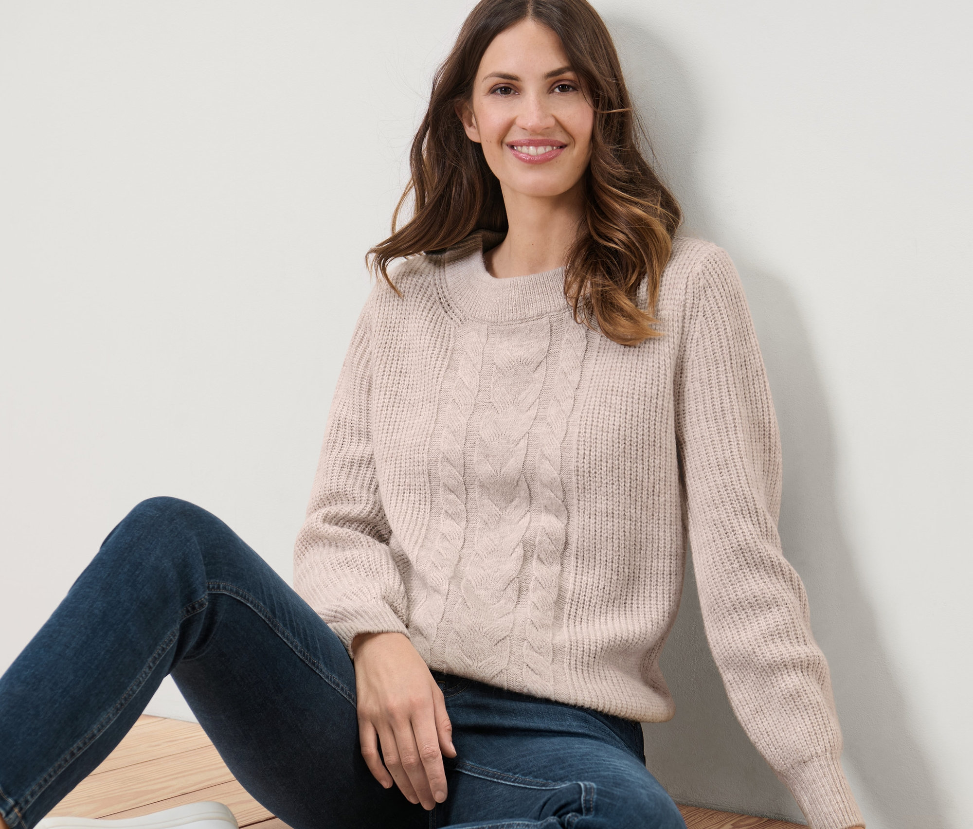 Žena sedí na podlaze a má na sobě svetr z hrubého úpletu s copánkovým vzorem.