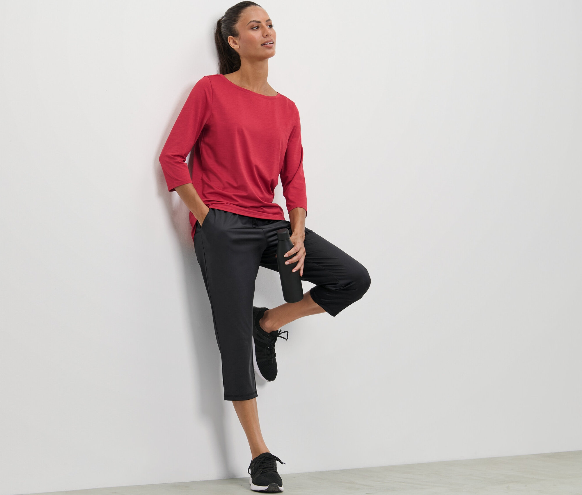 Žena opírající se o stěnu, má na sobě červené tričko a 3/4 sportovní kalhoty.