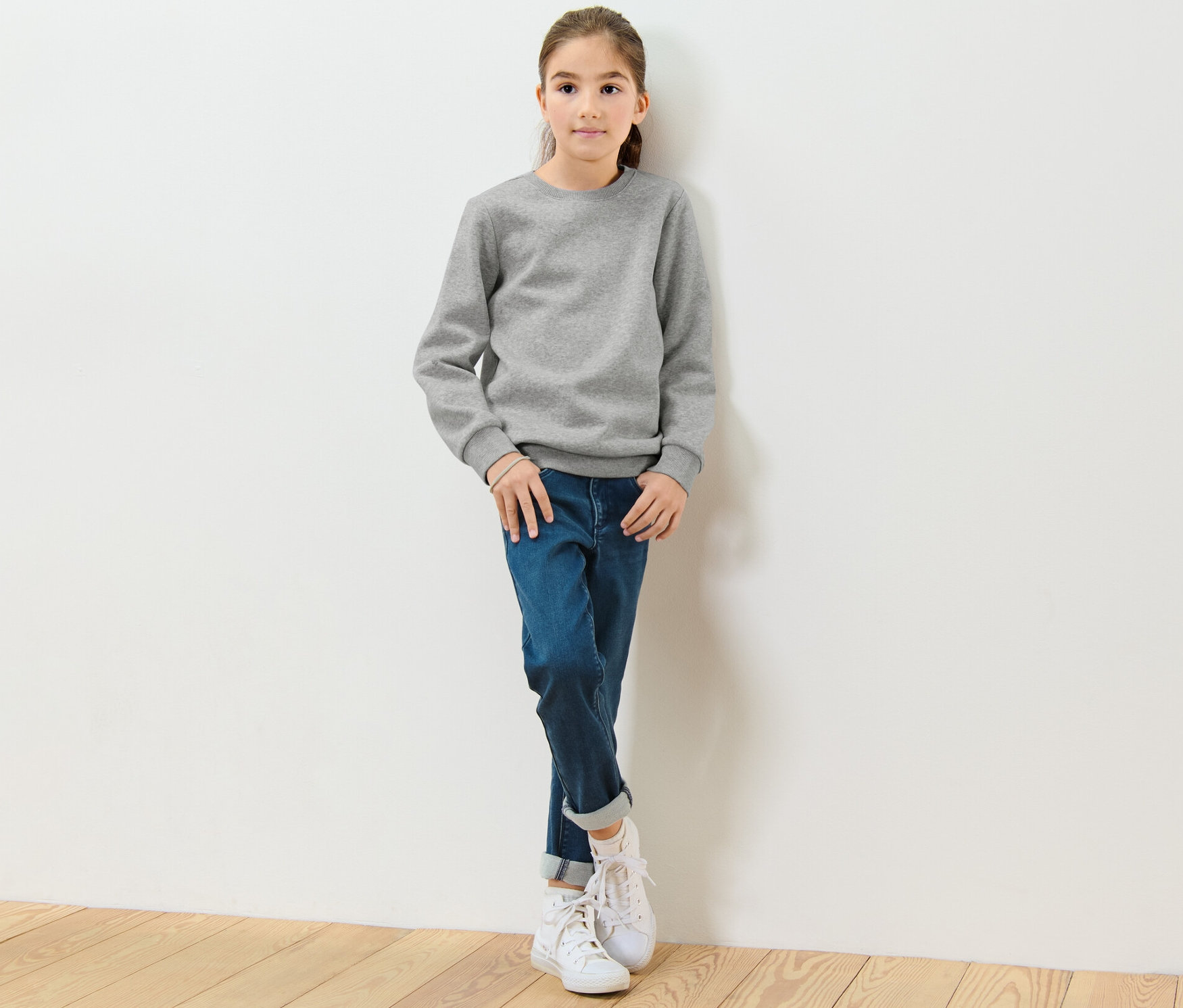 Dívka stojí opřená o bílou zeď a má na sobě šedou dětskou mikinu s počesanou vnitřní stranou.