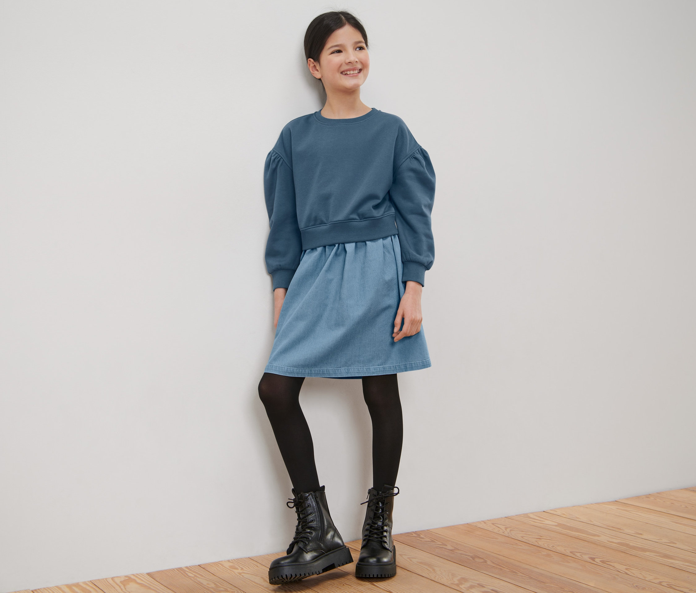 Usmívající se dívka v dívčích teplákových šatech stojí opřená o bílou zeď.