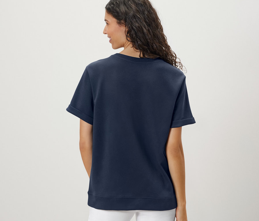 Zezadu je vidět žena s vlnitými vlasy, která má na sobě tmavě modré tričko z modalové směsi a bílé kalhoty.